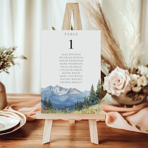 May include: A white table number card with the number 1 and a list of names, displayed on a small wooden easel. The card features a watercolor painting of mountains, trees, and wildflowers. The table setting includes plates, flowers, and decorative elements.