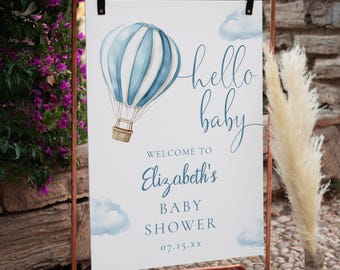 May include: A baby shower welcome sign featuring a blue and white hot air balloon watercolour design. The sign says "hello baby" and welcomes guests to Elizabeth's baby shower. It is held in a copper-coloured metal frame.