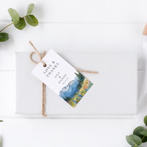 May include: A white gift box tied with twine, featuring a tag with the words "Love & Thanks" and the names "Lily & Daniel". The tag has a watercolor painting of a mountain landscape with blue, green, and yellow hues.