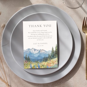 May include: A wedding thank you card with a watercolor mountain scene, placed on a gray plate with gold silverware. The card reads "THANK YOU" and includes a message from Lily & Daniel.