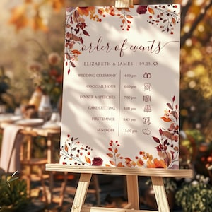 May include: A wooden easel displays a wedding sign with the words "Order of Events." The sign features a cream background with burgundy and orange floral accents. The schedule includes the wedding ceremony, cocktail hour, dinner, cake cutting, first dance, and send-off.