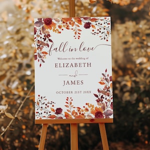 Puede incluir: Un letrero de bienvenida de boda blanco sobre un caballete de madera, con el texto "fall in love" en escritura cursiva. El letrero está decorado con elementos florales burdeos, naranjas y marrones. Los nombres "Elizabeth y James" se muestran, con la fecha "OCTOBER 21ST 20XX".