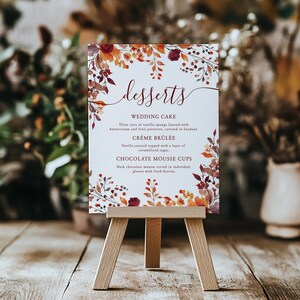 May include: A dessert menu displayed on a small wooden easel. The sign features the word "desserts" in elegant script, with a list of dessert options. The sign is decorated with floral elements in shades of orange, red, and brown.