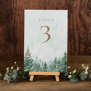 May include: A table number card with a watercolor forest scene. The card features the word "TABLE" and the number "3" in gold lettering. The card is displayed on a small wooden easel, with greenery in the foreground.