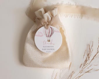 May include: A beige burlap gift bag with a round tag featuring a watercolor hot air balloon design in pink and mauve. The tag reads "thanks", "Elizabeth's Baby Shower", and the date.