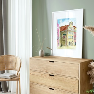 May include: A watercolor painting of a European city street scene with colorful buildings, a blue sky with clouds, and a cobblestone street. The painting is framed in a white frame and is hanging on a wall in a room with a wooden dresser and a chair.