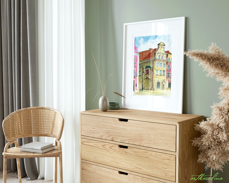 May include: A watercolor painting of a European city street scene with colorful buildings, a blue sky with clouds, and a cobblestone street. The painting is framed in a white frame and is hanging on a wall in a room with a wooden dresser and a chair.