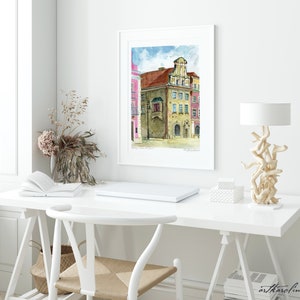 May include: A watercolor painting of a building with a balcony in a European city. The building is painted in shades of brown, yellow, and red. The sky is blue with white clouds. The painting is framed in a white frame and is hanging on a white wall.