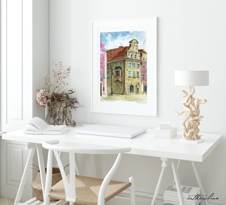 May include: A watercolor painting of a building with a balcony in a European city. The building is painted in shades of brown, yellow, and red. The sky is blue with white clouds. The painting is framed in a white frame and is hanging on a white wall.