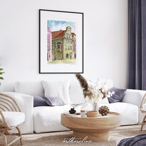 May include: A watercolor painting of a building with a pink facade and a yellow facade. The building has a large window and a balcony. The painting is framed in a black frame and is hanging on a white wall.
