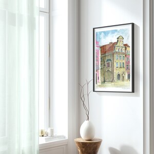May include: Watercolor painting of a colorful building with a unique architectural design. The building is painted in shades of pink, yellow, and brown, with a white balcony and a decorative roof. The painting is framed in black and has a white mat.
