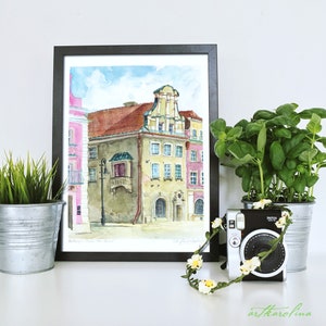 May include: A watercolor painting of a building with a pink facade and a green balcony. The painting is framed in black and is hanging on a white wall. There is a potted plant and a camera with a flower garland in front of the painting.