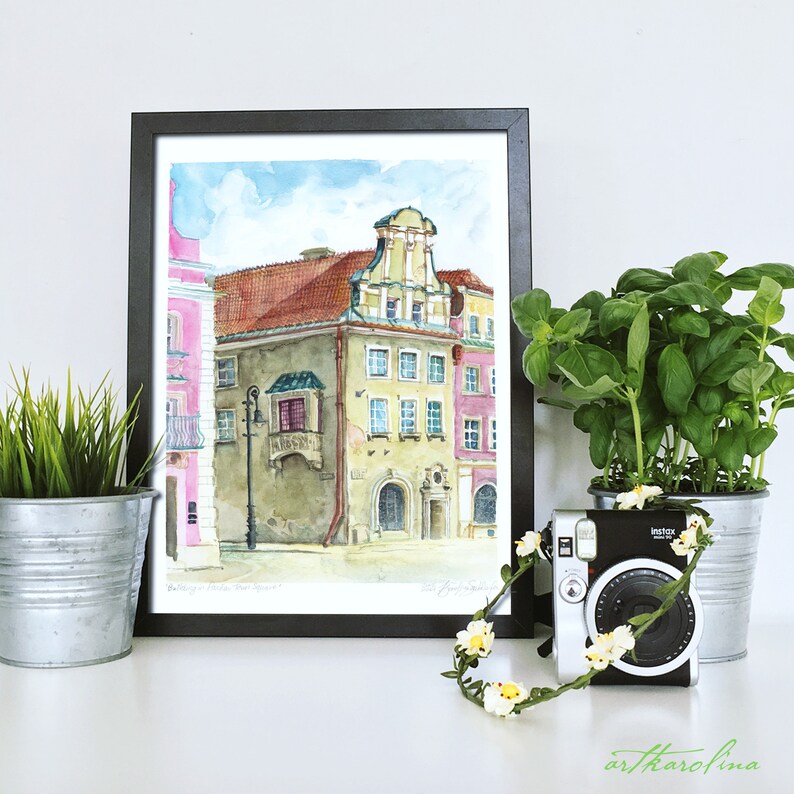 May include: A watercolor painting of a building with a pink facade and a green balcony. The painting is framed in black and is hanging on a white wall. There is a potted plant and a camera with a flower garland in front of the painting.