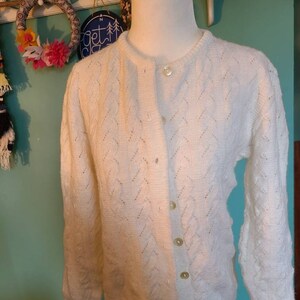 May include: A white knitted cardigan with a button-up front and a diamond pattern. The cardigan has long sleeves and a relaxed fit.