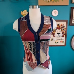 May include: A sleeveless button-front vest featuring a patchwork design of various knit fabrics in shades of red, blue, beige, and black. The garment has a deep V-neckline, dark blue ribbed trim along the armholes and front, and four buttons.