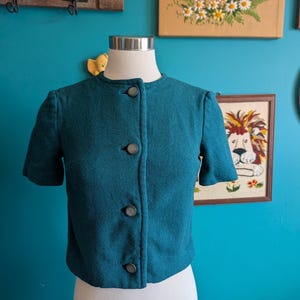 May include: A short-sleeved, teal-colored jacket featuring a round neckline and a front button closure with four dark, circular buttons. The garment is constructed from a textured, woven fabric and is displayed on a white mannequin torso.