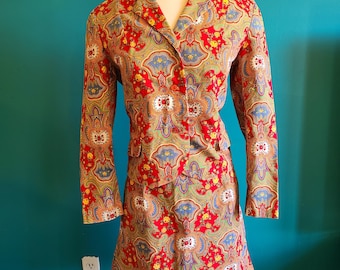 May include: A red, blue, and gold paisley print skirt suit. The skirt is a-line and the jacket has a collar and long sleeves.