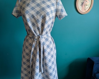 May include: A vintage blue and white patterned dress with short sleeves and a matching tie belt. The dress is displayed on a mannequin. The dress has a high neckline and a diamond pattern.