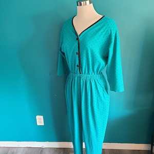 May include: A teal blue jumpsuit with black polka dots and a black trim around the neckline. The jumpsuit has a button closure at the front and a drawstring waist.