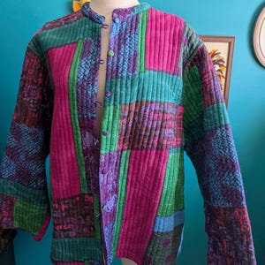 May include: A quilted jacket in a patchwork design, featuring vibrant blocks of pink, green, purple, and blue. The jacket has a mandarin collar and is fastened with small, round buttons. The fabric has a textured, quilted appearance.
