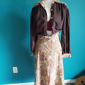 K&ouml;nnte beinhalten: Eine braune Bluse mit langen &Auml;rmeln, einem Kragen mit Blumendruck und einem passenden Blumenrock. Die Bluse hat einen Knopfleistenverschluss und der Rock einen G&uuml;rtel.
