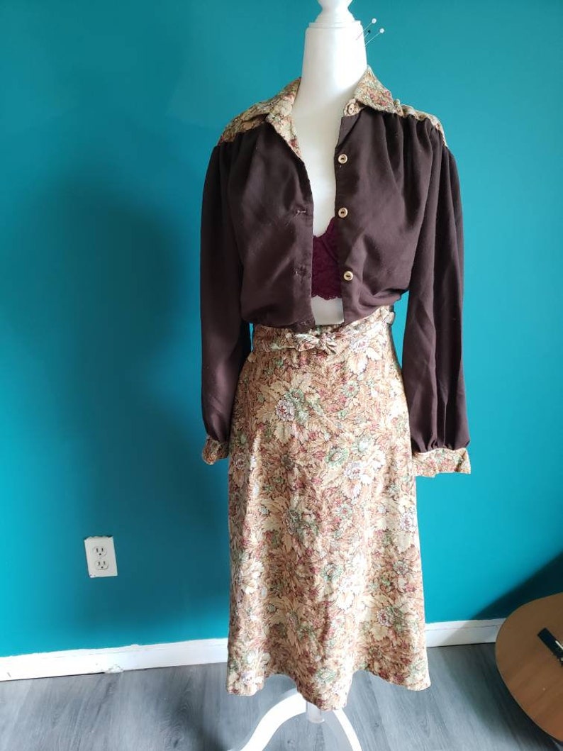 K&ouml;nnte beinhalten: Eine braune Bluse mit langen &Auml;rmeln, einem Kragen mit Blumendruck und einem passenden Blumenrock. Die Bluse hat einen Knopfleistenverschluss und der Rock einen G&uuml;rtel.