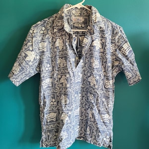 May include: A blue short-sleeved button-down shirt with a white floral pattern. The shirt has a collar and a pocket on the left chest. The label on the shirt reads "Reyn Spooner".
