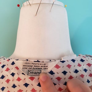 May include: A white blouse with a pink and blue geometric pattern. The blouse has a label that reads "100% Polyester, Machine Wash Warm, Line or Tumble Dry, Sears".