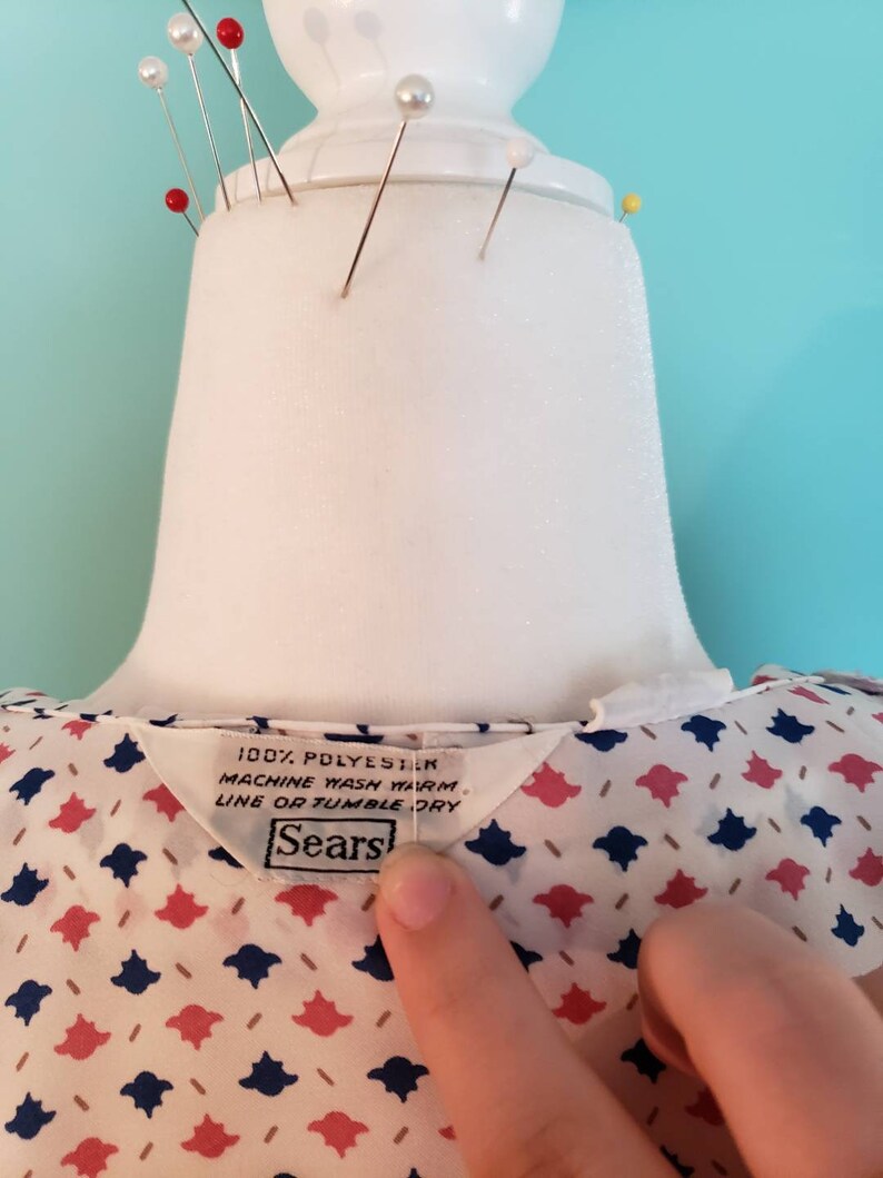 May include: A white blouse with a pink and blue geometric pattern. The blouse has a label that reads "100% Polyester, Machine Wash Warm, Line or Tumble Dry, Sears".