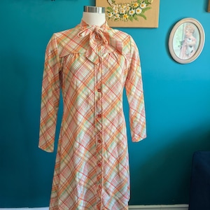 May include: A vintage, long-sleeved dress in a plaid pattern of orange, red, yellow, and white. The dress features a bow at the neck and a button-down front. The dress is displayed on a mannequin.
