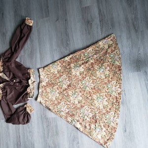 K&ouml;nnte beinhalten: Eine braune Bluse mit langen &Auml;rmeln und einem Rock mit Blumenmuster. Die Bluse hat einen Knopfleistenverschluss und einen gerafften Ausschnitt. Der Rock ist A-f&ouml;rmig und hat eine hohe Taille.