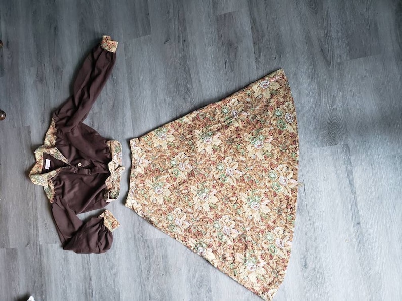 K&ouml;nnte beinhalten: Eine braune Bluse mit langen &Auml;rmeln und einem Rock mit Blumenmuster. Die Bluse hat einen Knopfleistenverschluss und einen gerafften Ausschnitt. Der Rock ist A-f&ouml;rmig und hat eine hohe Taille.