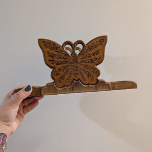 May include: A brown wooden napkin holder featuring a butterfly-shaped cutout with intricate, engraved scrollwork patterns on the wings. The butterfly is centered on a horizontal base, showcasing a natural wood grain finish and detailed carvings.