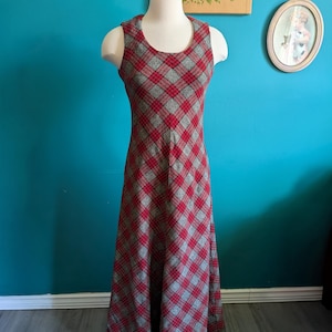 May include: A sleeveless, floor-length dress on a mannequin. The dress features a red and gray plaid pattern. The dress has a scoop neckline and a flared skirt. The mannequin is set against a teal wall.