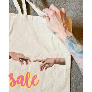 Könnte beinhalten: Eine beige Canvas-Tragetasche mit einem Aufdruck von zwei Händen, die sich gegenseitig erreichen. Das Wort "Sale" ist in rosa Buchstaben mit gelbem Umriss auf der Tasche geschrieben.