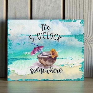 May include: A rectangular sign featuring a watercolour beach scene with a coconut drink, a small umbrella, and a yellow flower. The text reads It's 5 O'CLOCK somewhere in black script and print lettering against a blue and white ocean background.