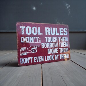 A distressed red metal sign reading TOOL RULES standing upright on a gray wooden surface against a dark gray wall.