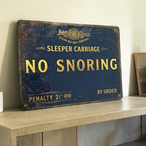 May include: A rectangular dark blue metal sign with distressed edges and gold lettering. It features a steam train logo, the text "STEAM RAILWAY COMPANY", "SLEEPER CARRIAGE", "NO SNORING", "BY ORDER", and "PENALTY 2 1/2d" with two circular coin icons.