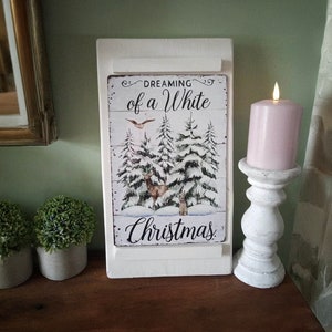 May include: A white wooden sign with the text "Dreaming of a White Christmas" and a winter scene with deer and snow-covered trees. A lit pink candle sits on a white candlestick holder. Two small potted plants are on the left.