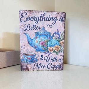 May include: A rectangular metal sign with a distressed finish featuring a blue teapot and teacup illustration, floral accents, and the text Everything is Better With a Nice Cuppa in dark blue script lettering against a light pink background.