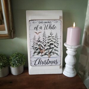 May include: A white wooden sign with the text "Dreaming of a White Christmas" and a winter scene with deer and snow-covered trees. A lit pink candle sits on a white candlestick holder. Two small potted plants are on the left.