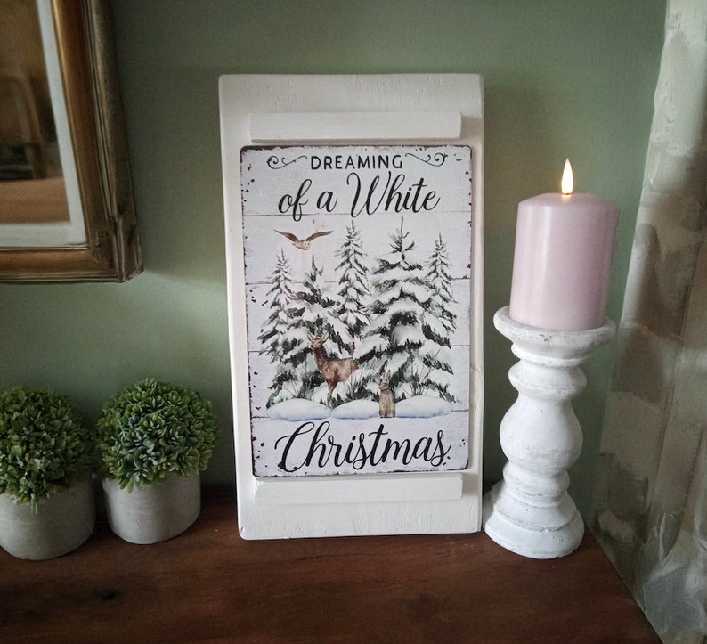 May include: A white wooden sign with the text "Dreaming of a White Christmas" and a winter scene with deer and snow-covered trees. A lit pink candle sits on a white candlestick holder. Two small potted plants are on the left.