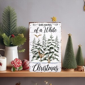 May include: A wooden sign with the text "Dreaming of a White Christmas" and a winter scene with deer and snow-covered trees. The sign is displayed on a wooden shelf with Christmas decorations, including candles, small Christmas trees, and greenery.