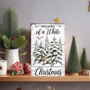 May include: A wooden sign with the text "Dreaming of a White Christmas" features a winter scene with snow-covered trees and deer. The sign is displayed on a wooden shelf with Christmas decorations, including candles, small Christmas trees, and greenery.