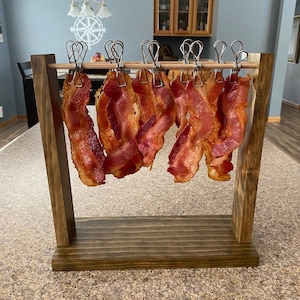 May include: A wooden bacon rack with several strips of cooked bacon hanging from metal clips. The rack is stained a dark brown and has a rectangular base. The bacon is a deep red-brown color, with crispy edges.