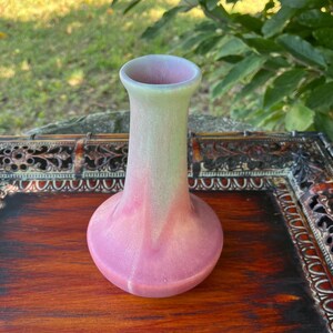 May include: A small, pink and green glazed ceramic vase with a flared base and a narrow neck. The vase is sitting on a wooden tray with intricate carvings.
