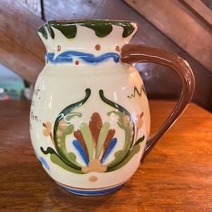 May include: Cream-colored ceramic pitcher with a brown handle and decorative floral design. The pitcher features a green rim, blue accents, and a central motif of stylized leaves in green, blue, and brown. The pitcher has text on the side.