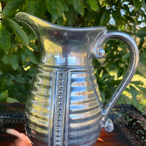 May include: A silver-colored pitcher with a curved handle and a spout. The pitcher has a ribbed body with vertical panels adorned with small, round details. The pitcher is sitting on a wooden surface.