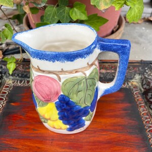 May include: A white ceramic pitcher with a blue handle and rim. The pitcher is decorated with a hand-painted design of fruit, including grapes, apples, and pears.