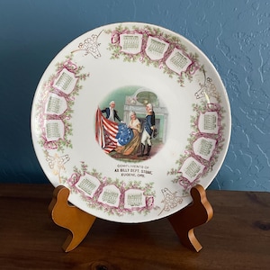 May include: A white decorative plate with a gold rim, featuring a central illustration of historical figures and an American flag. The plate is adorned with calendar designs and the text "COMPLIMENTS OF AX BILLY DEPT. STORE, EUGENE, ORE."